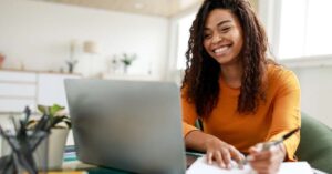 Image of a smiling woman writing on a piece of paper and using a laptop