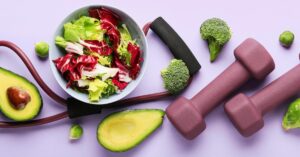 Image of a bowl of cabbage and lettuce on a table with a cut avocado, hand weights, and pieces of broccoli