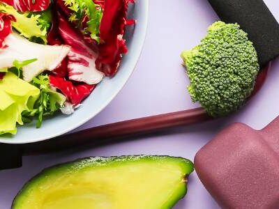 Image of a bowl of cabbage and lettuce on a table with a cut avocado, hand weights, and pieces of broccoli