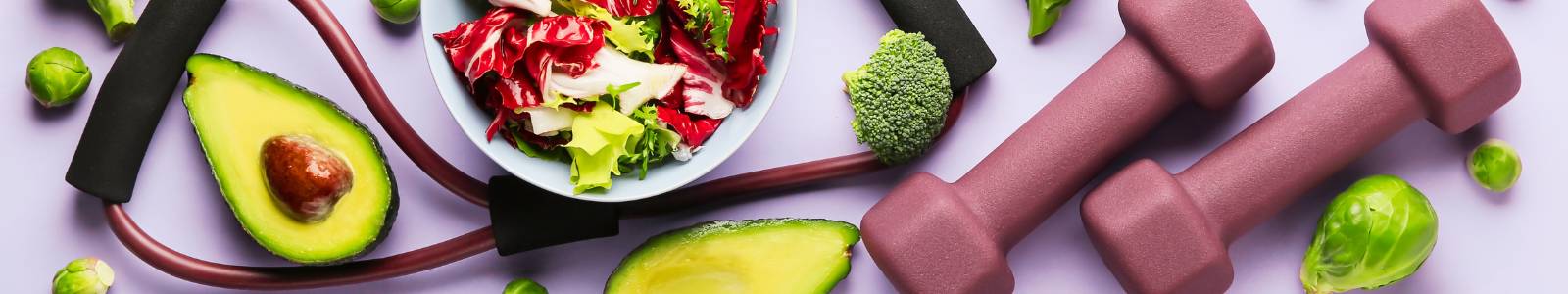 Image of a bowl of cabbage and lettuce on a table with a cut avocado, hand weights, and pieces of broccoli