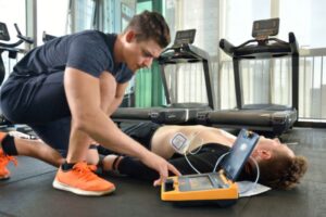 Man helping another man with an AED in a gym setting.